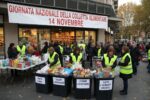 Volontari con pettorali gialli organizzano la raccolta alimentare fuori da un negozio con un grande striscione rosso.